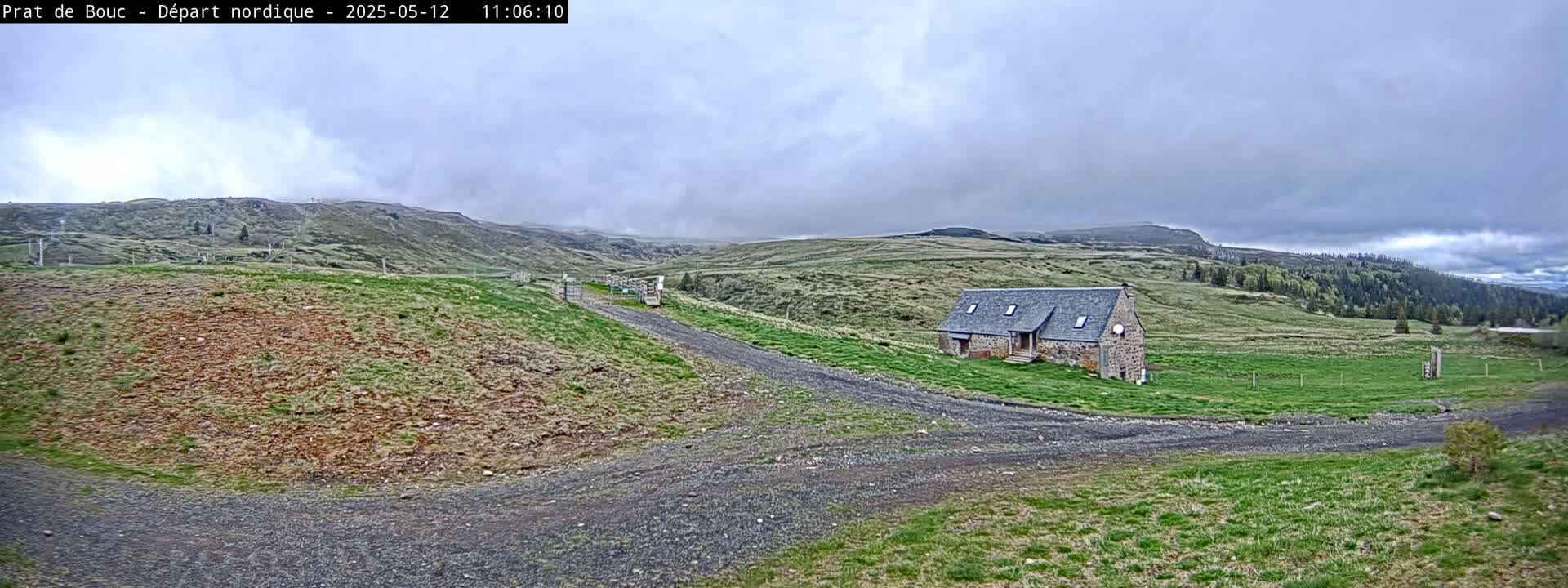 A stone building sits on a grassy hillside, with a gravel road leading to it under an overcast sky.