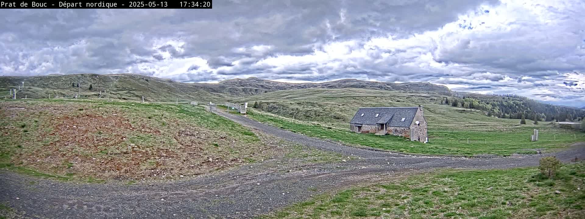 A stone building sits on a grassy hillside with a gravel road leading to it under a partly cloudy sky.