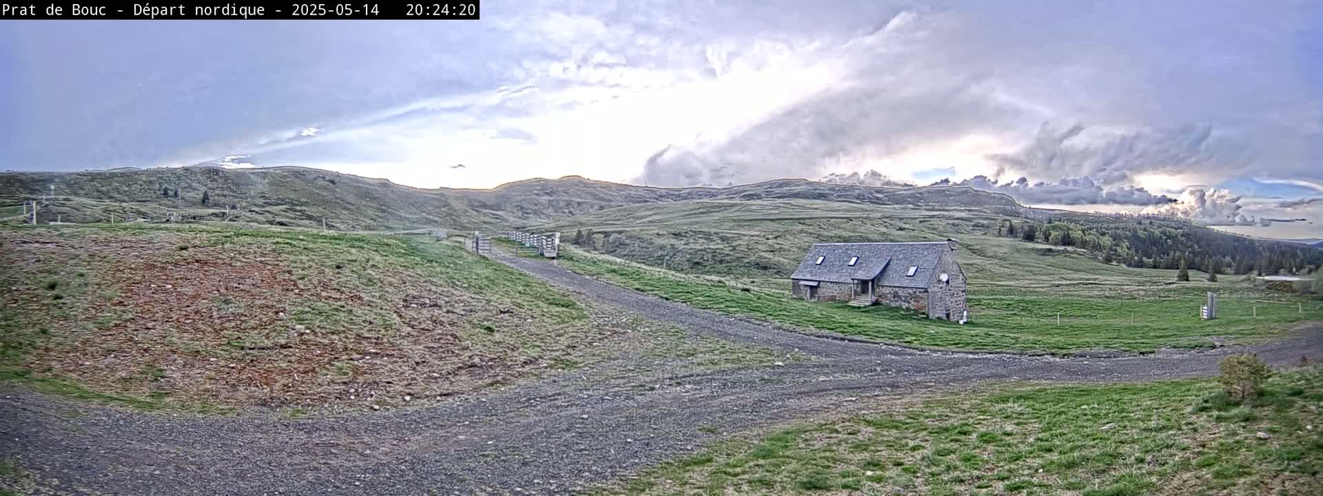 A stone house sits on a grassy hillside under a partly cloudy sky, with a gravel road leading to it.