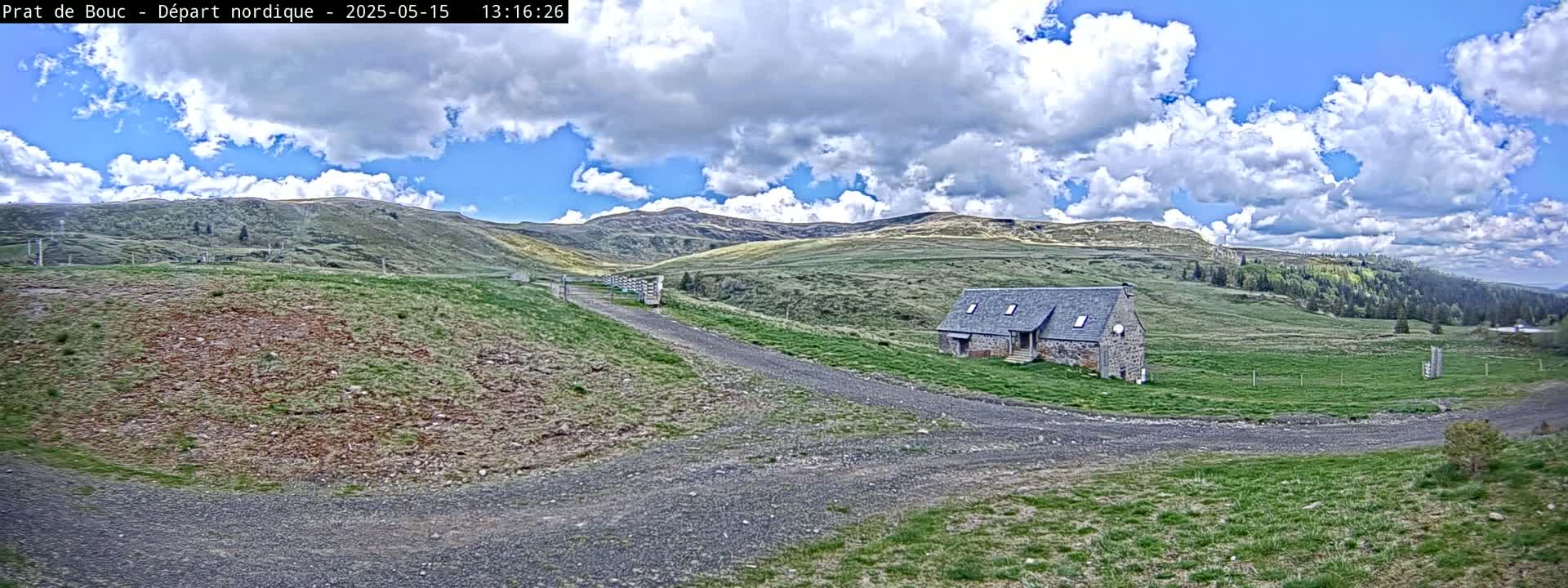 A stone building sits on a grassy hilltop beneath a partly cloudy blue sky, with gravel roads leading to it.