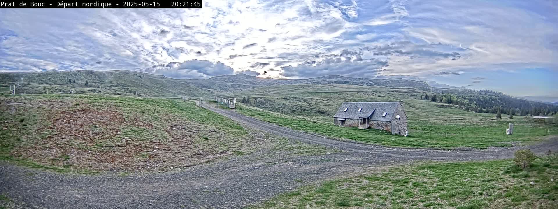 A stone house sits on a grassy hill overlooking a gravel road, under a partly cloudy sky.