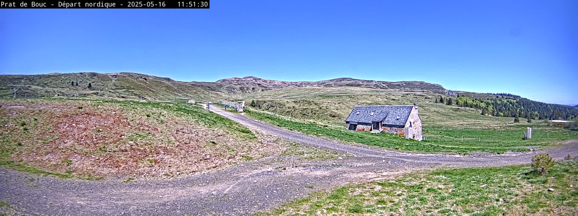 A gravel road branches near a stone building in a grassy, hilly landscape under a clear blue sky.