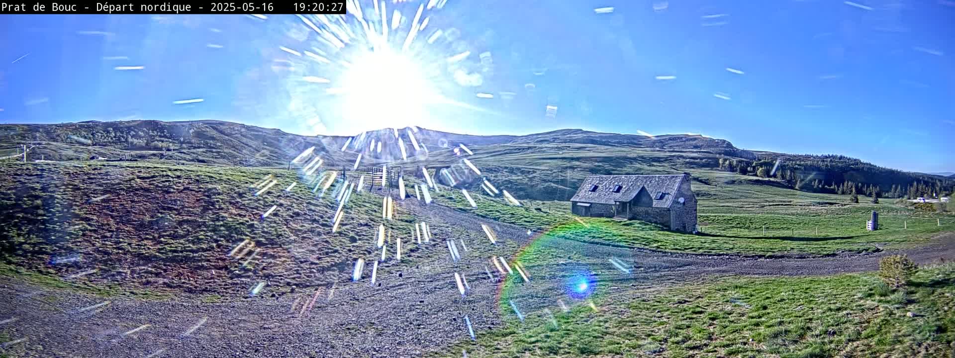 A stone building sits on a grassy hillside under a bright, sunny sky with some lens flare.