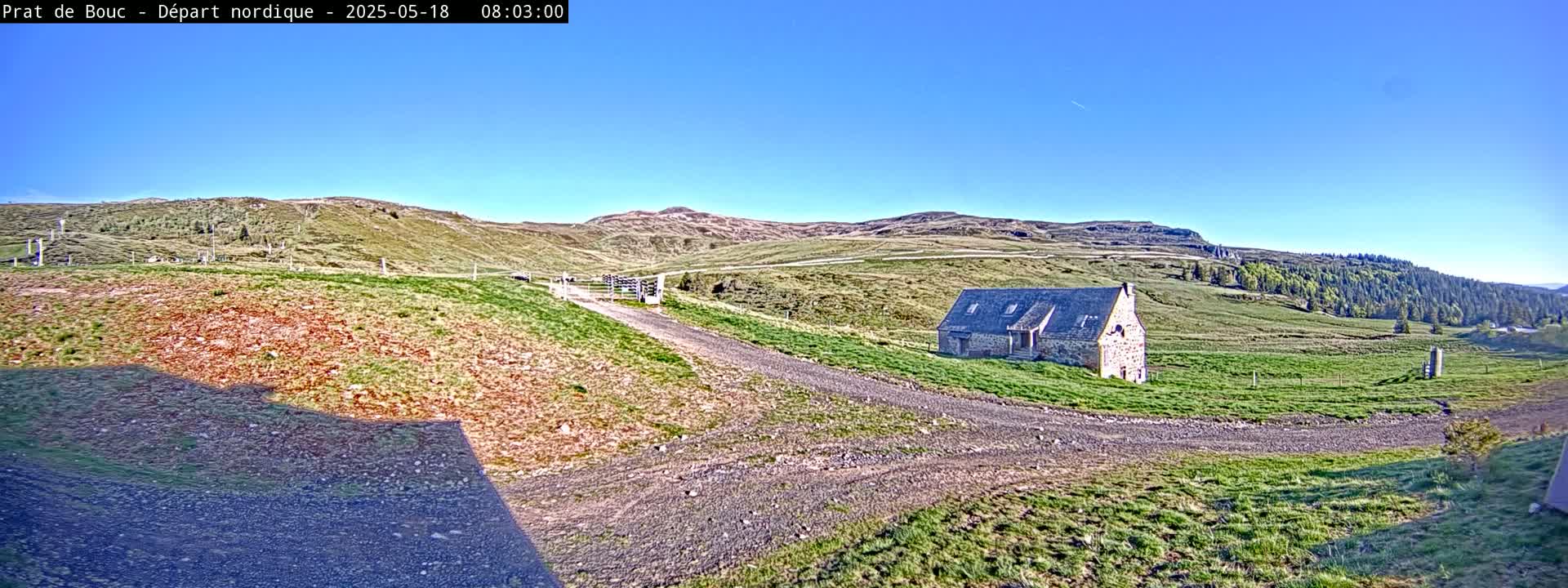 A gravel road leads to a stone building nestled in a grassy valley under a clear blue sky.