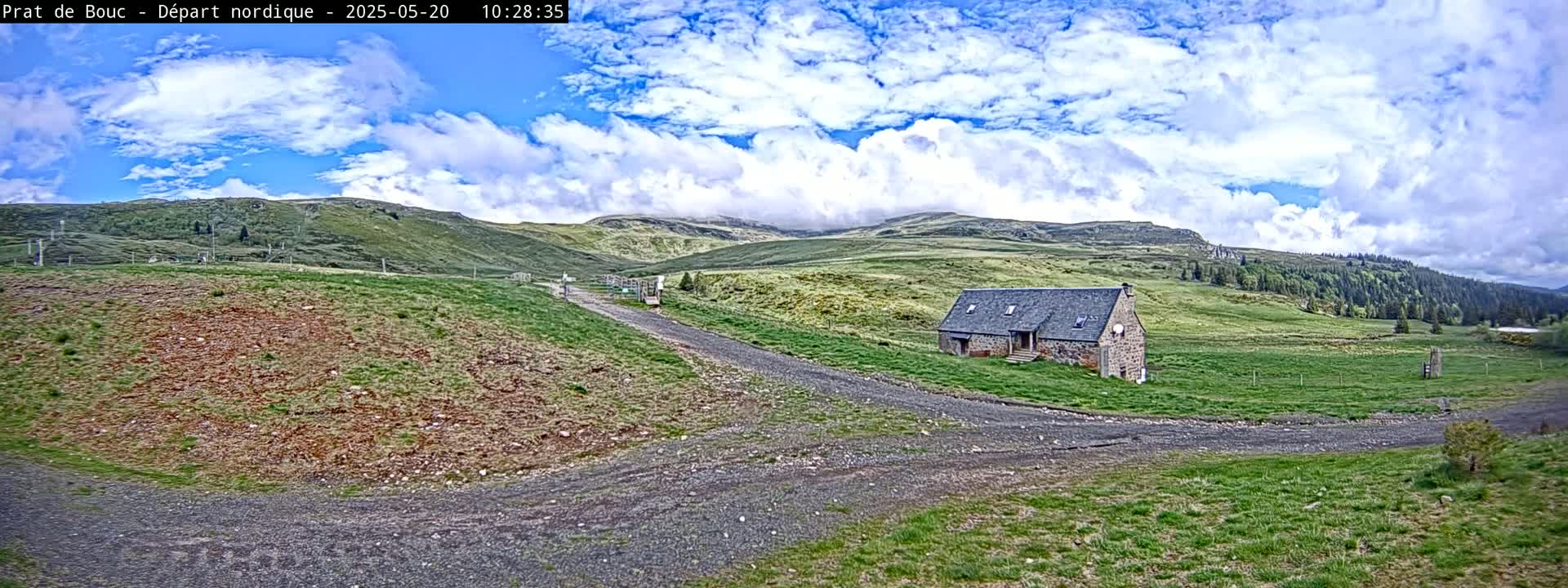 A stone building sits on a grassy hill overlooking a gravel road, under a partly cloudy sky.