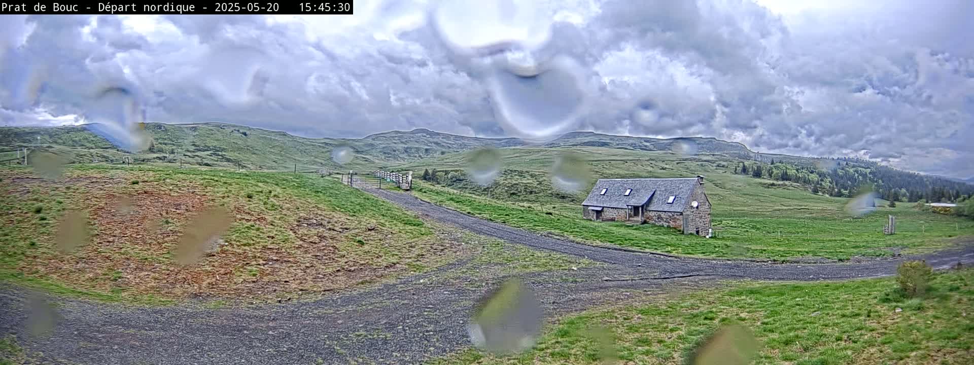 A stone house sits on a grassy hillside under a cloudy, rainy sky, with a gravel road leading to it.