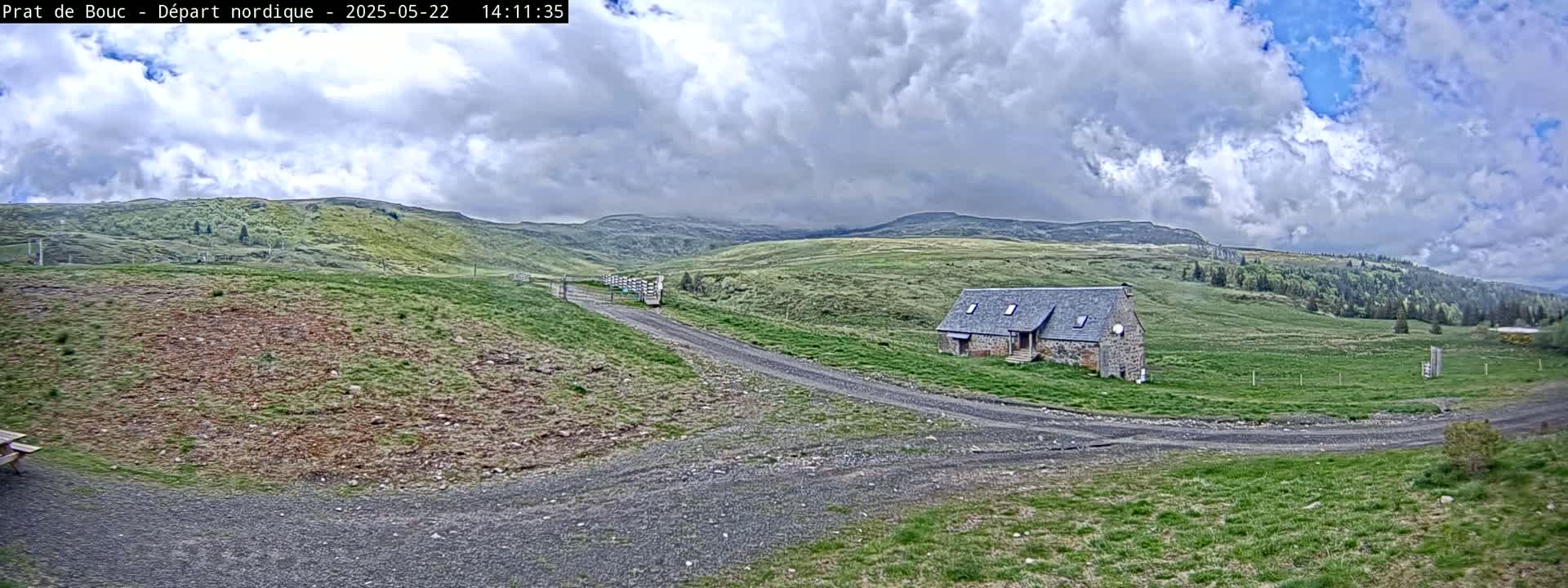 A stone building sits on a grassy hillside under a partly cloudy sky, with a dirt road leading toward it.