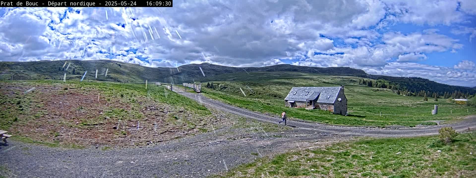 A person walks on a gravel path toward a stone building in a grassy, hilly landscape under a partly cloudy sky with light rain or snow falling.