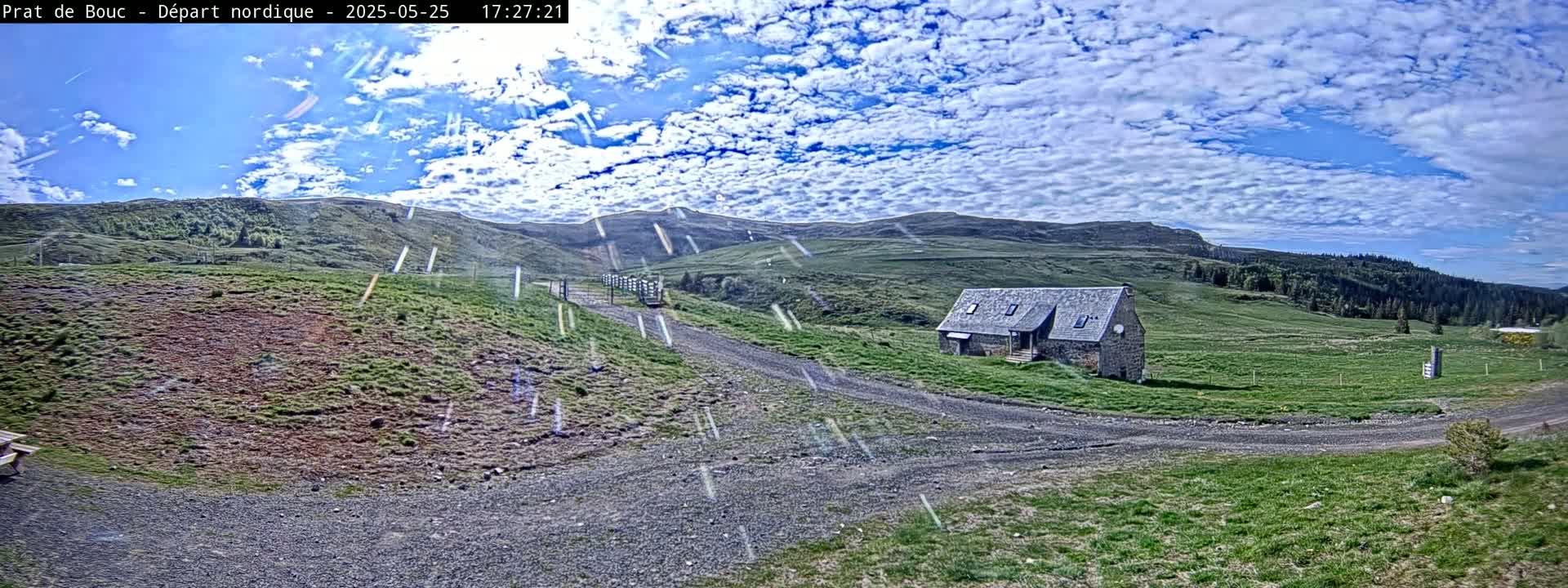 A stone house sits on a grassy hillside under a partly cloudy sky with light rain or snow falling.