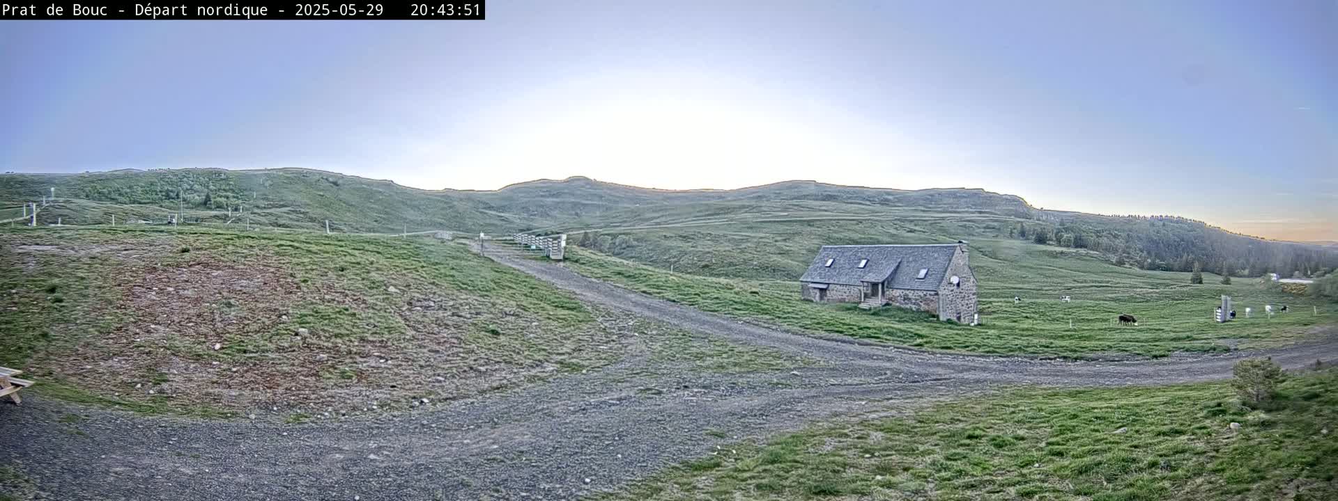 A stone building sits on a grassy hillside under a clear, twilight sky, with a gravel road leading to it and livestock grazing in a nearby field.