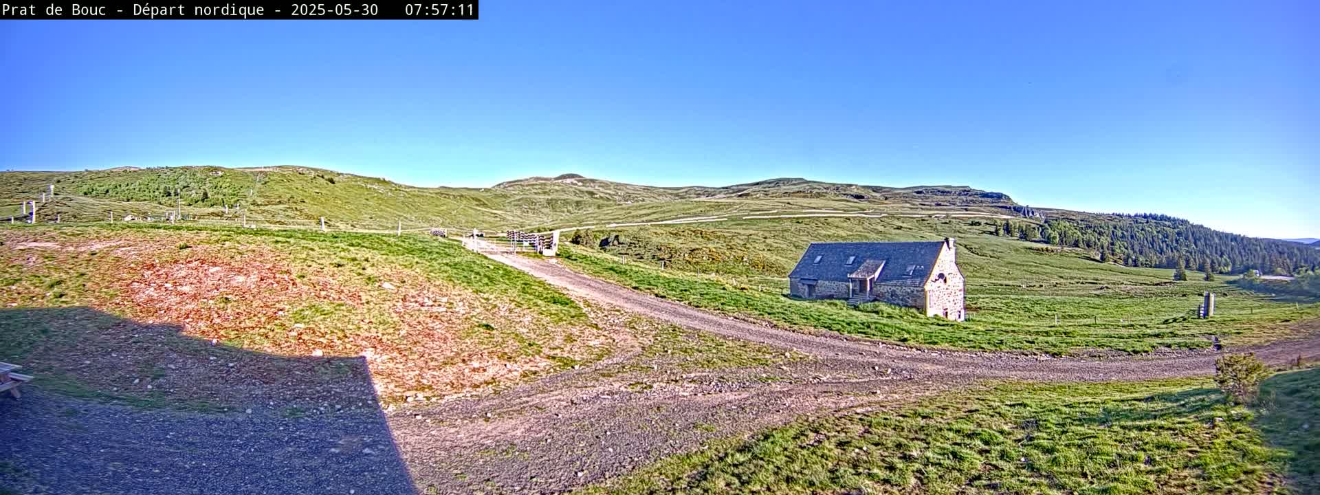 A stone building sits on a grassy hillside under a clear blue sky, with a gravel road leading to it.