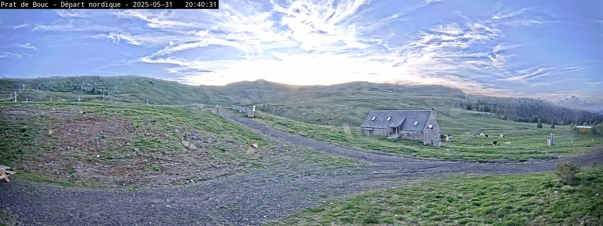 A stone building sits on a grassy hillside under a partly cloudy sunset sky, with a gravel road leading to it and cattle grazing in a nearby field.