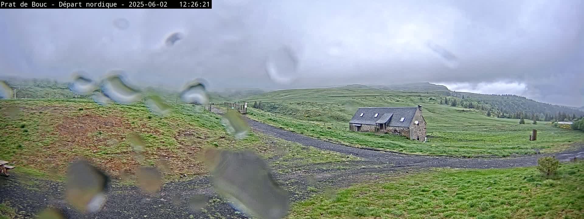 Rain falls on a grassy landscape with a stone building and a gravel road, all under a cloudy sky.