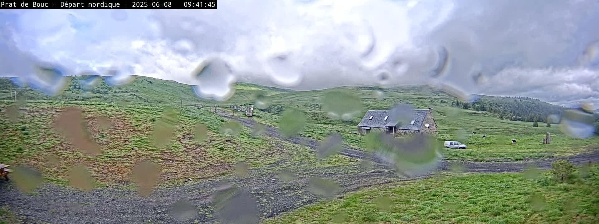 A stone house sits on a grassy hillside under a cloudy, rainy sky, with a few cows grazing in a nearby field.
