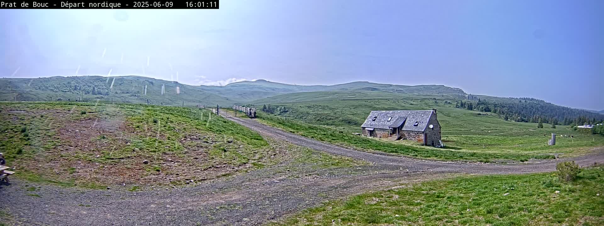 A stone building sits on a grassy hilltop with a gravel road leading to it, under a clear, sunny sky.