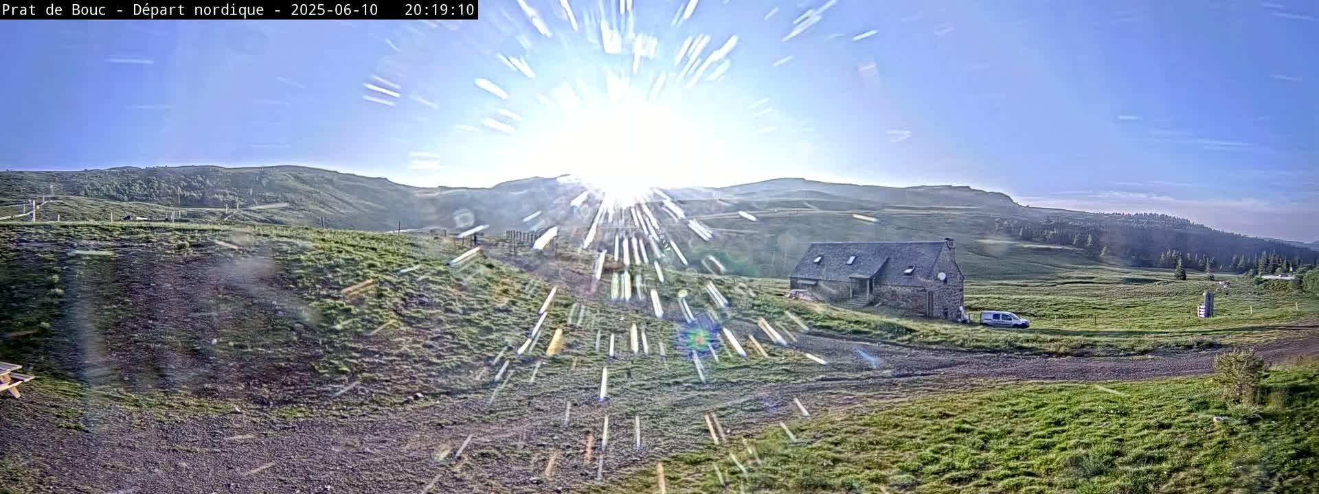 A bright sun shines over a grassy hillside with a stone building and a vehicle parked nearby, under a clear blue sky.