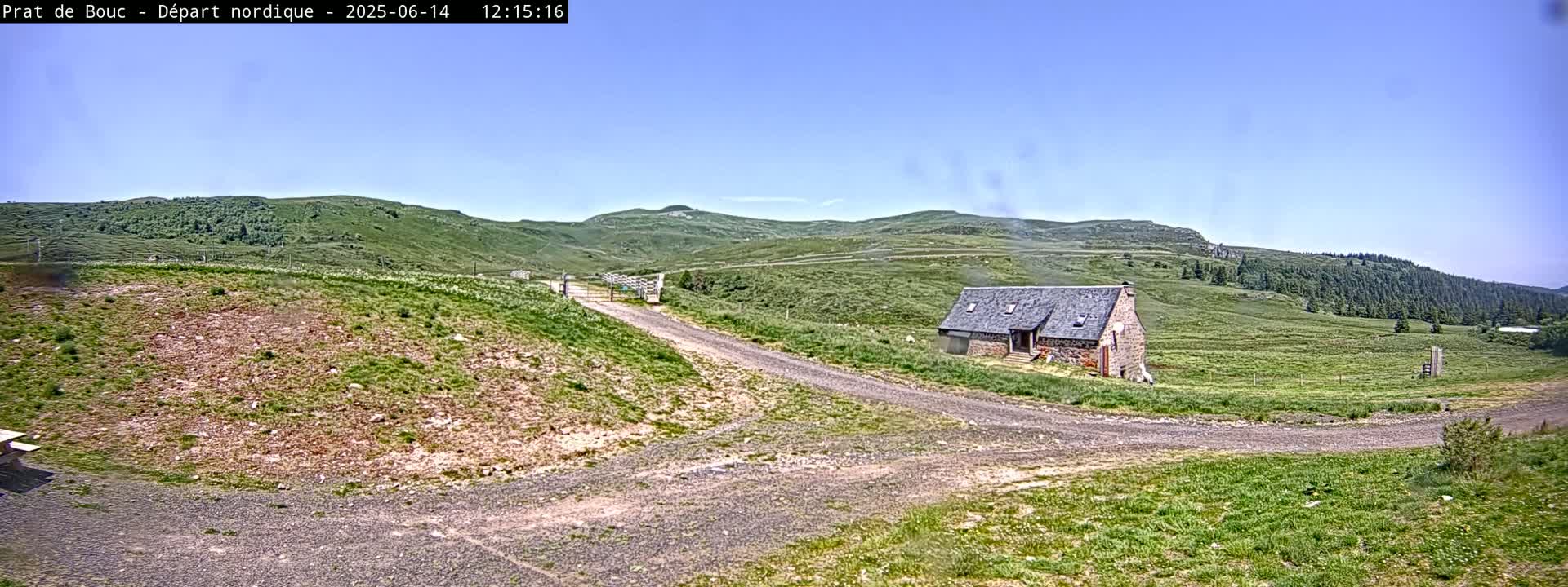 A stone building sits on a grassy hillside next to a dirt road under a clear blue sky.