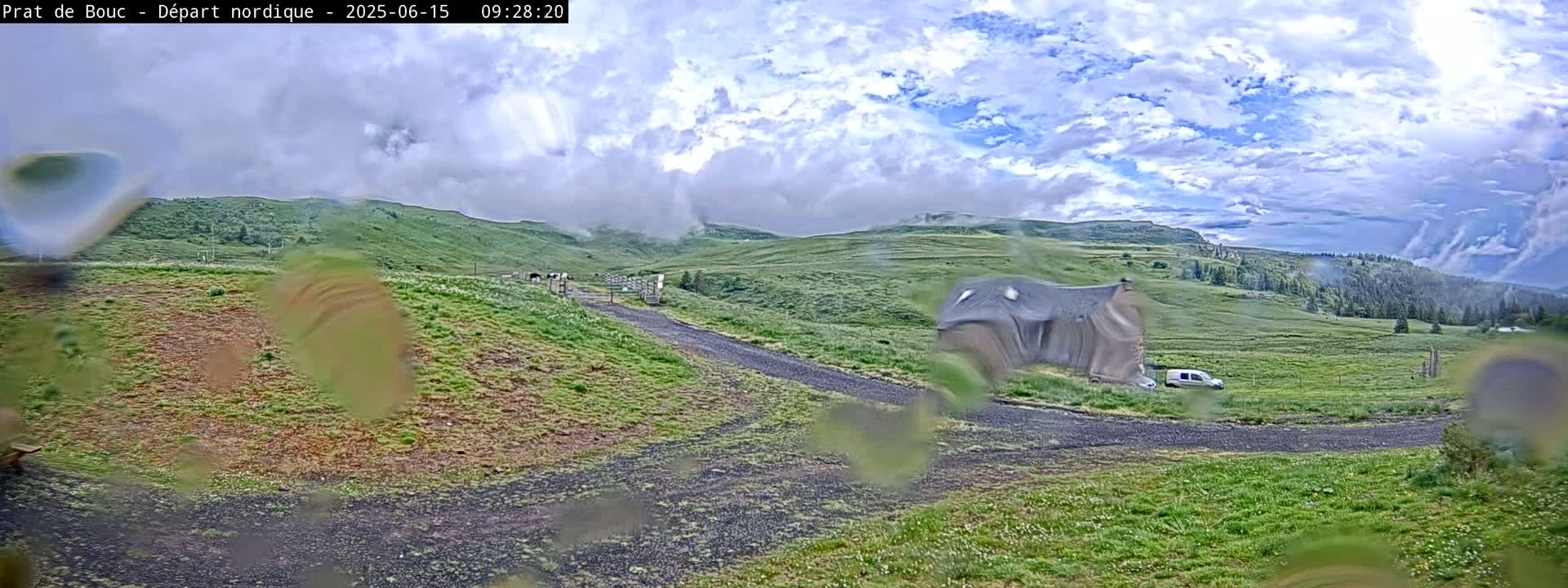 A partly cloudy sky overlooks a grassy hillside with a building, a vehicle, and a few cows visible through rain-covered glass.