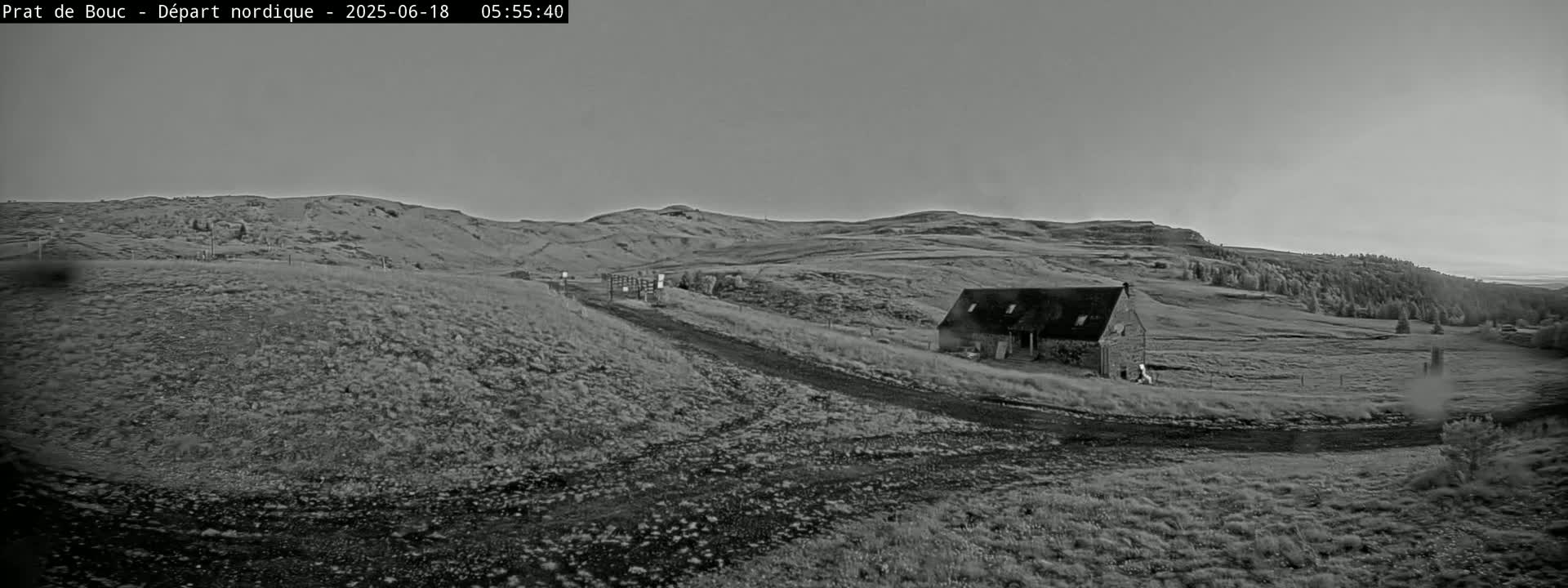 A grayscale image shows a long, low building situated on a grassy hillside under a clear, pale sky.