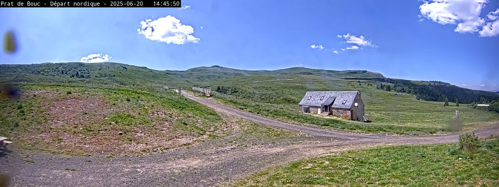 A stone building sits on a grassy hillside with a dirt road leading to it under a mostly sunny sky with some fluffy clouds.
