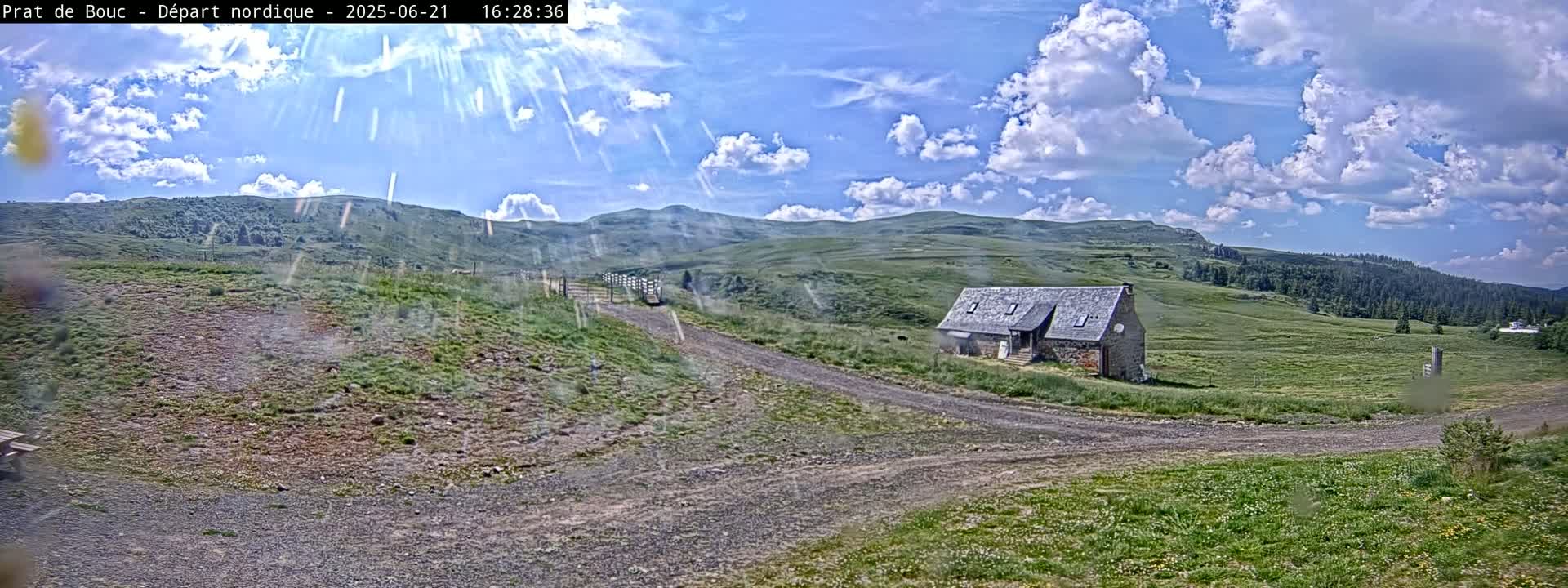 A stone building sits on a grassy hillside under a partly cloudy, sunny sky, with dirt roads leading to it.