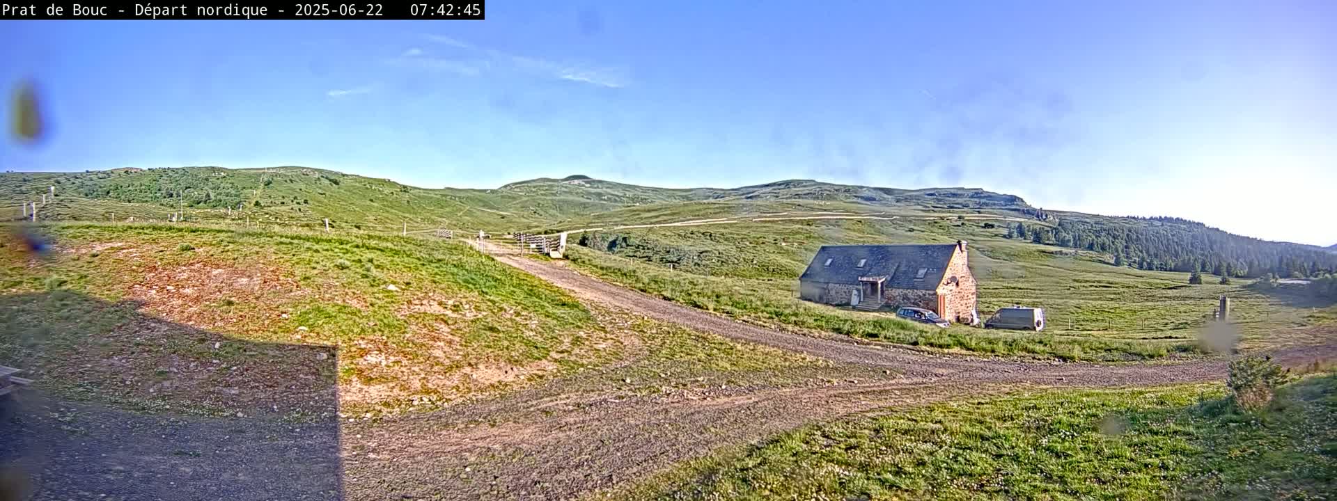 A stone building sits on a grassy hillside next to a dirt road under a clear blue sky.
