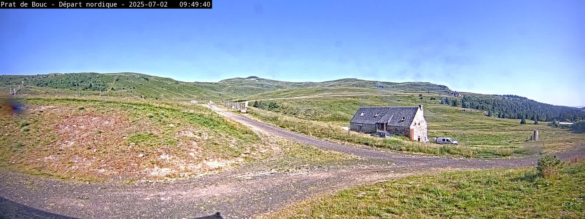 A stone building sits beside a gravel road on a grassy hillside under a clear blue sky.