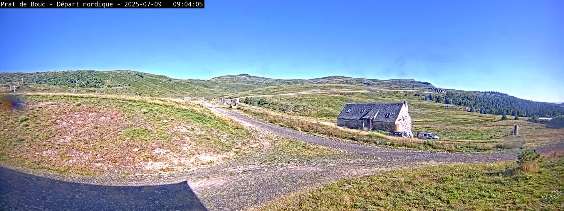 A stone house sits on a grassy hillside near a gravel road under a clear blue sky.