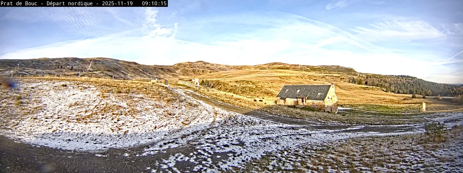 A wide panoramic view captures a vast, rolling landscape covered in dry golden grass and patches of thin snow, with a stone house and winding dirt path under a bright, partly cloudy sky.