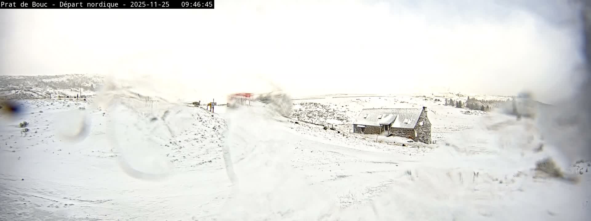 A blustery winter scene unfolds across a snow-covered mountainous landscape featuring a stone building, distant ski infrastructure, and visible blowing snow under bright, overcast skies, partially obscured by moisture on the camera lens.