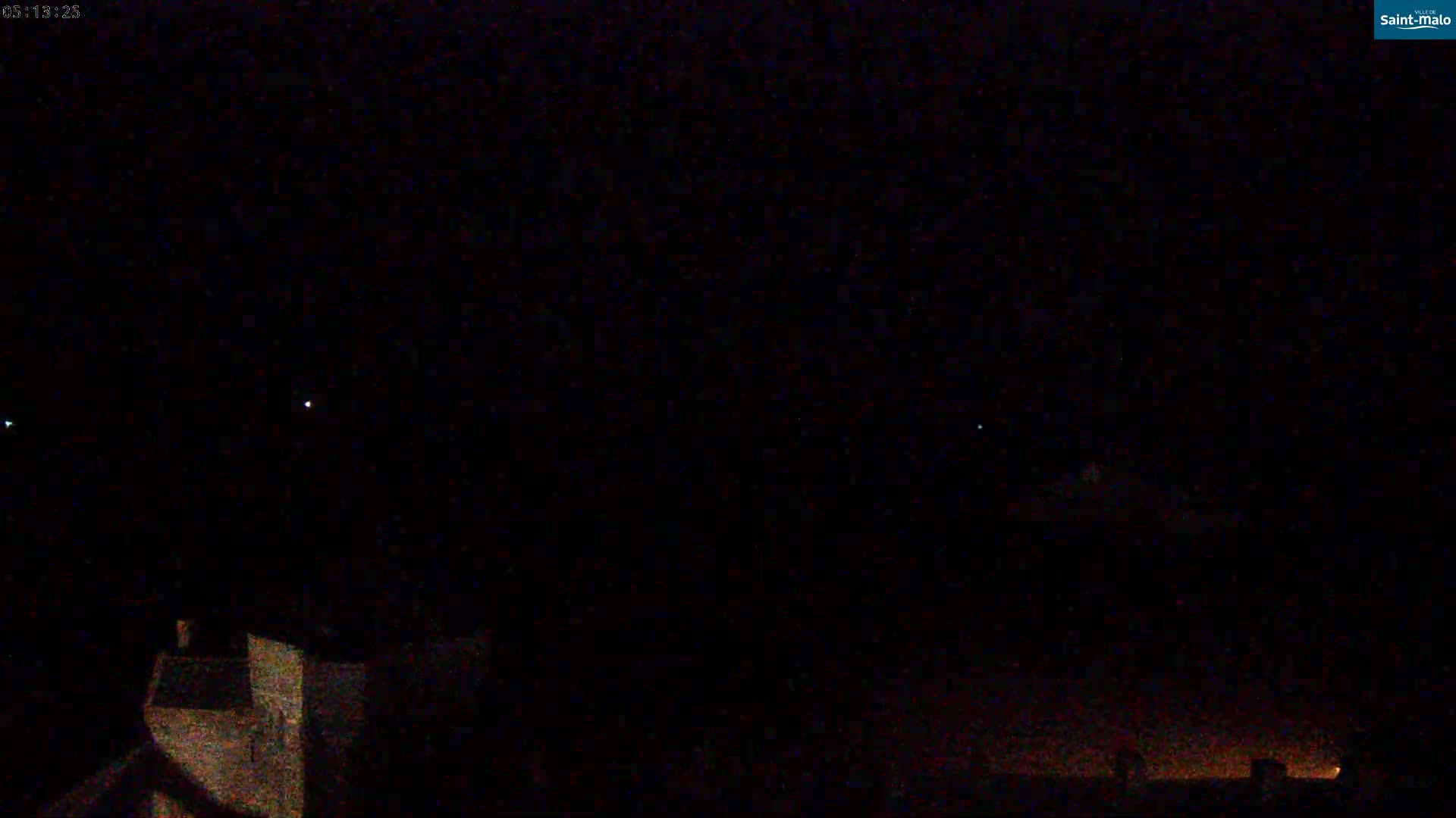 A nighttime view of a dark sky with a few faint lights visible, possibly stars, and the silhouettes of buildings in the foreground.