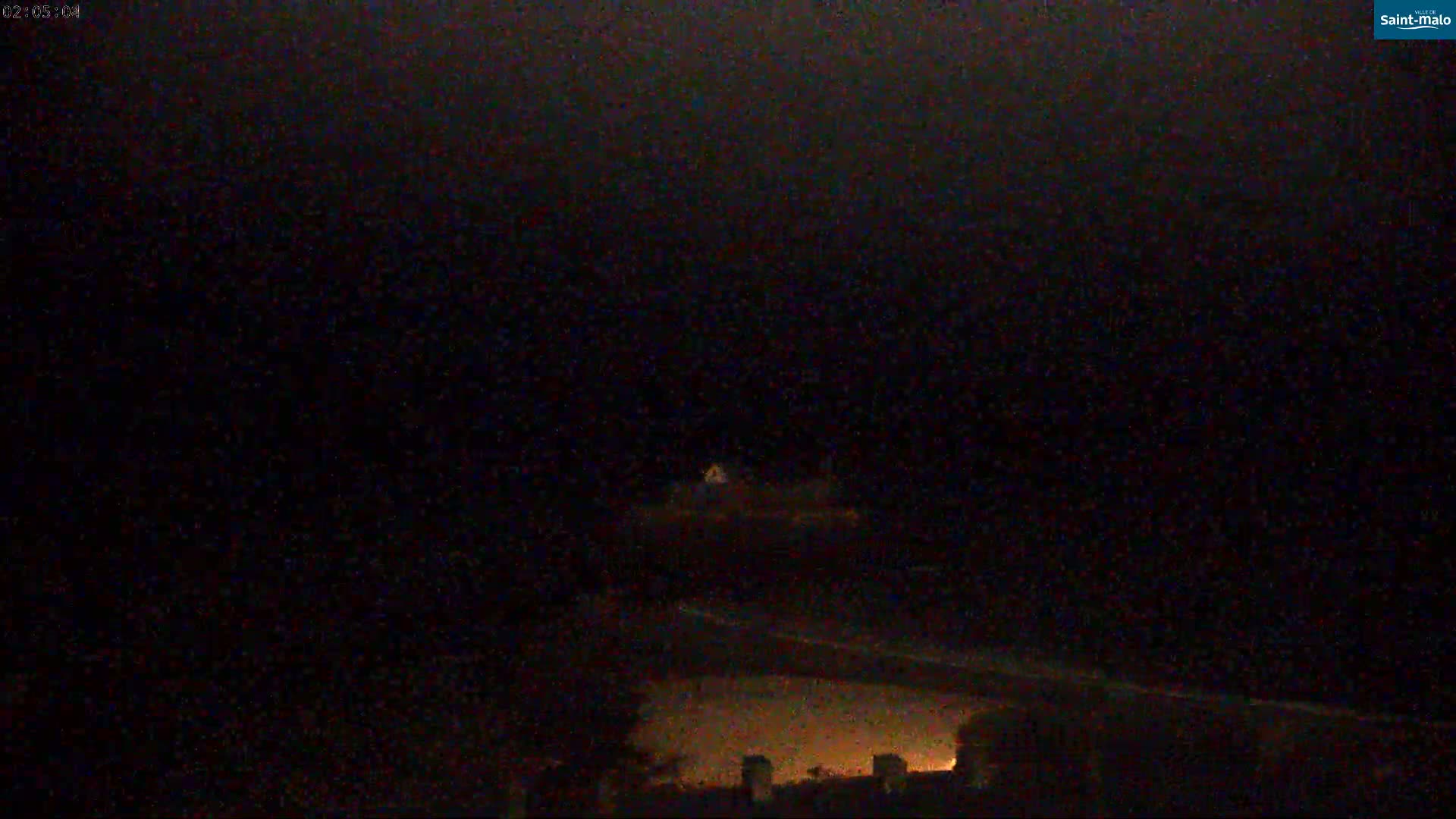 A dark, nighttime scene shows a dimly lit landscape with a few lights visible in the distance under an overcast sky.