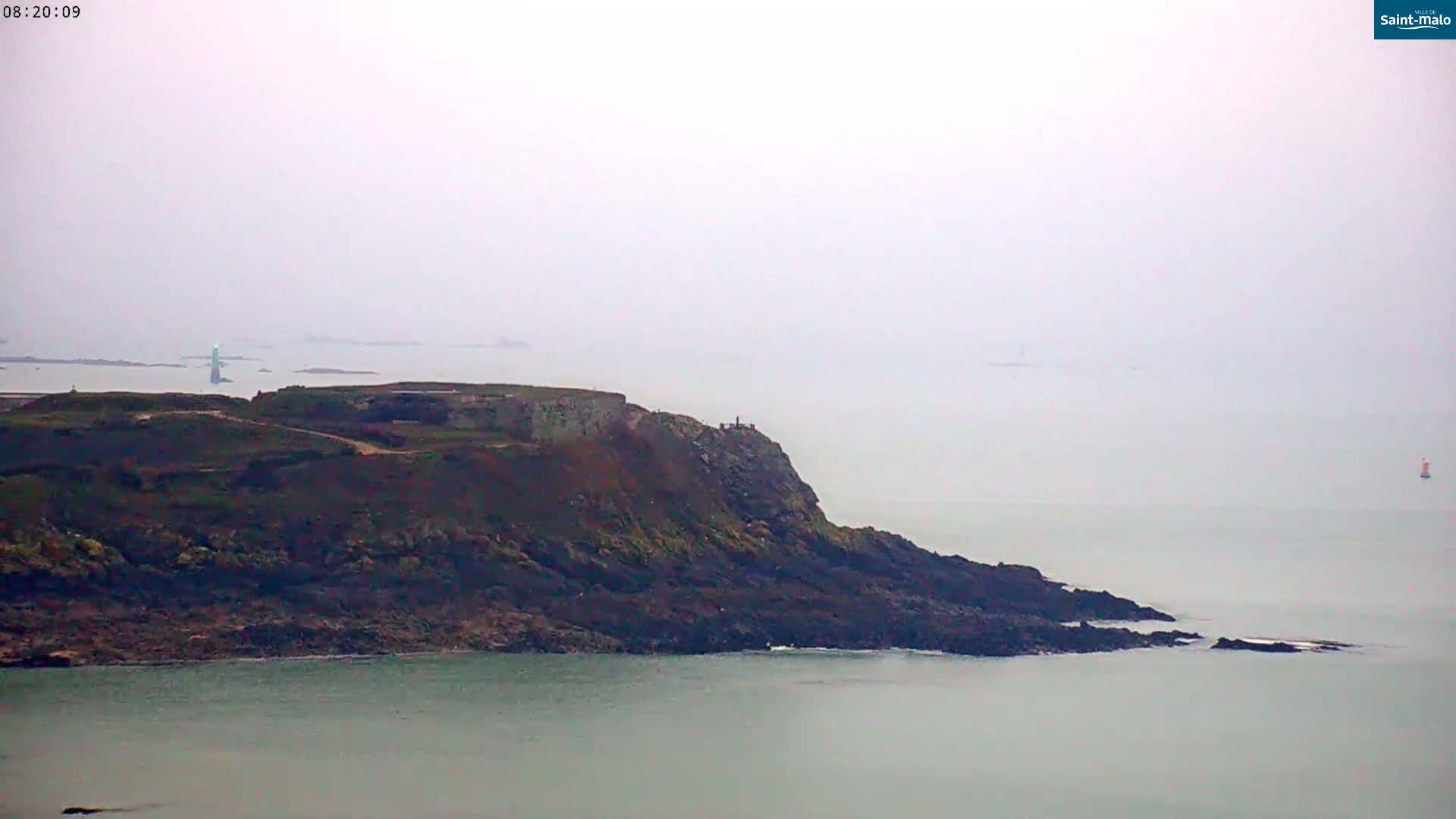 A rocky, dark-colored headland extends into a calm, foggy sea under a hazy sky.