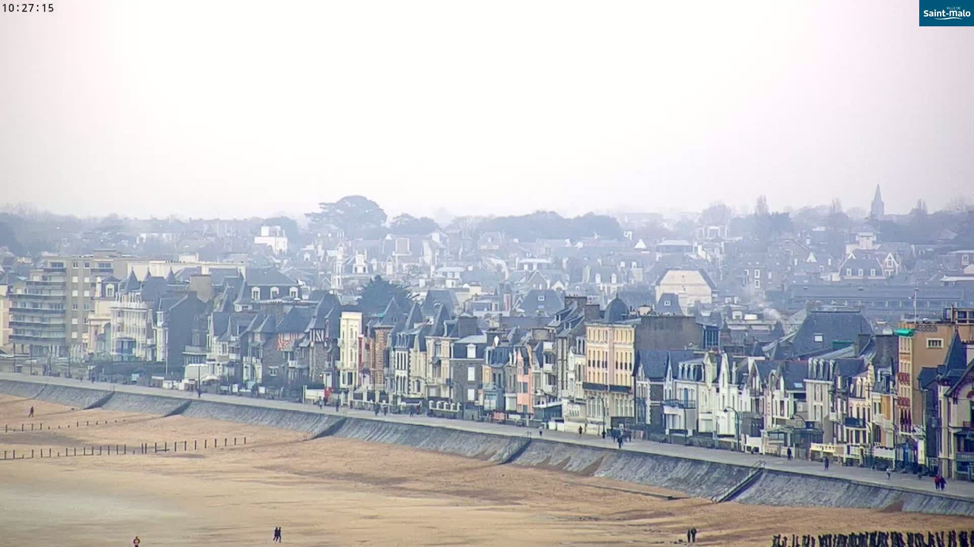 A hazy coastal town with many closely-packed buildings stretches along a sandy beach, backed by a larger expanse of town.