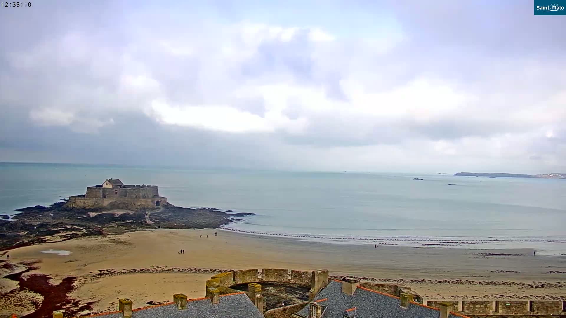 A coastal scene shows a stone castle on a rocky outcrop next to a sandy beach with a calm sea under a cloudy sky.