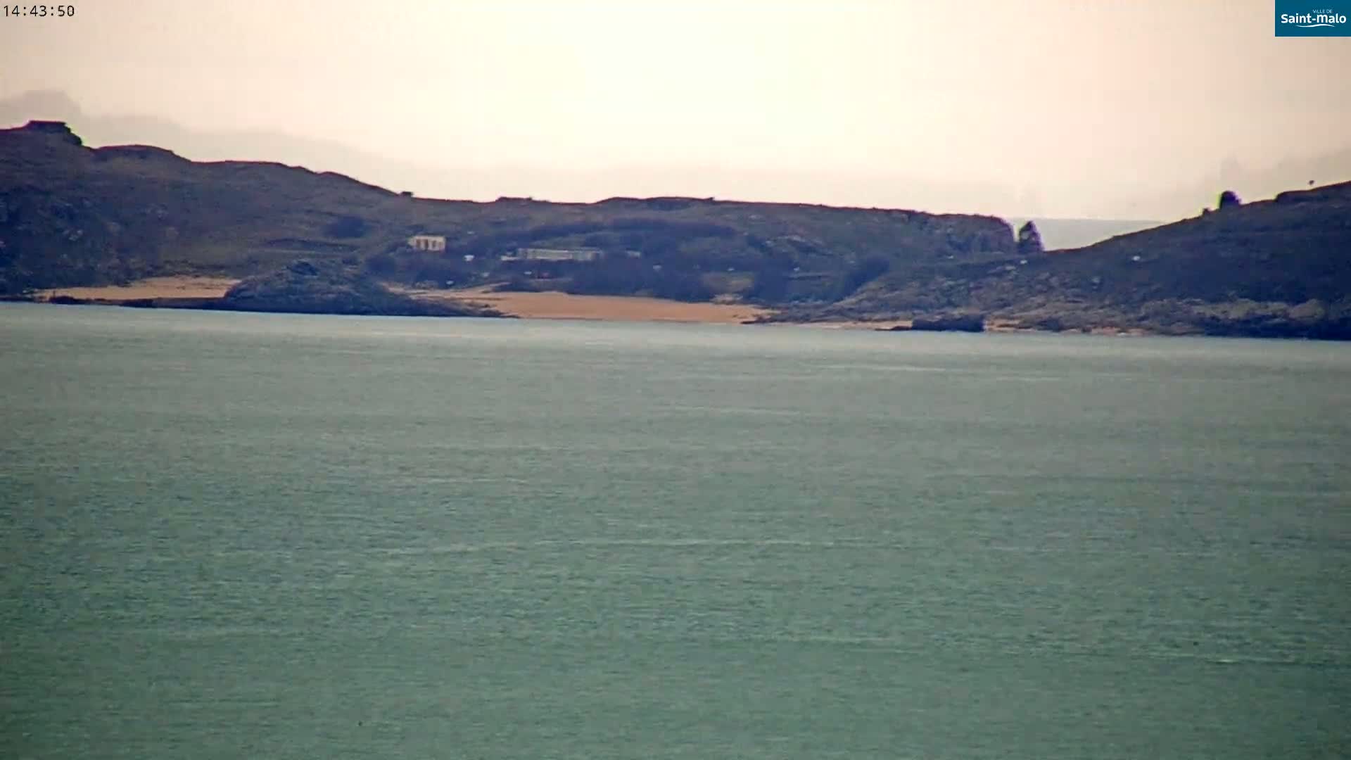 A calm, gray-toned body of water stretches out to a distant shoreline featuring dark hills, a small sandy beach, and some buildings.