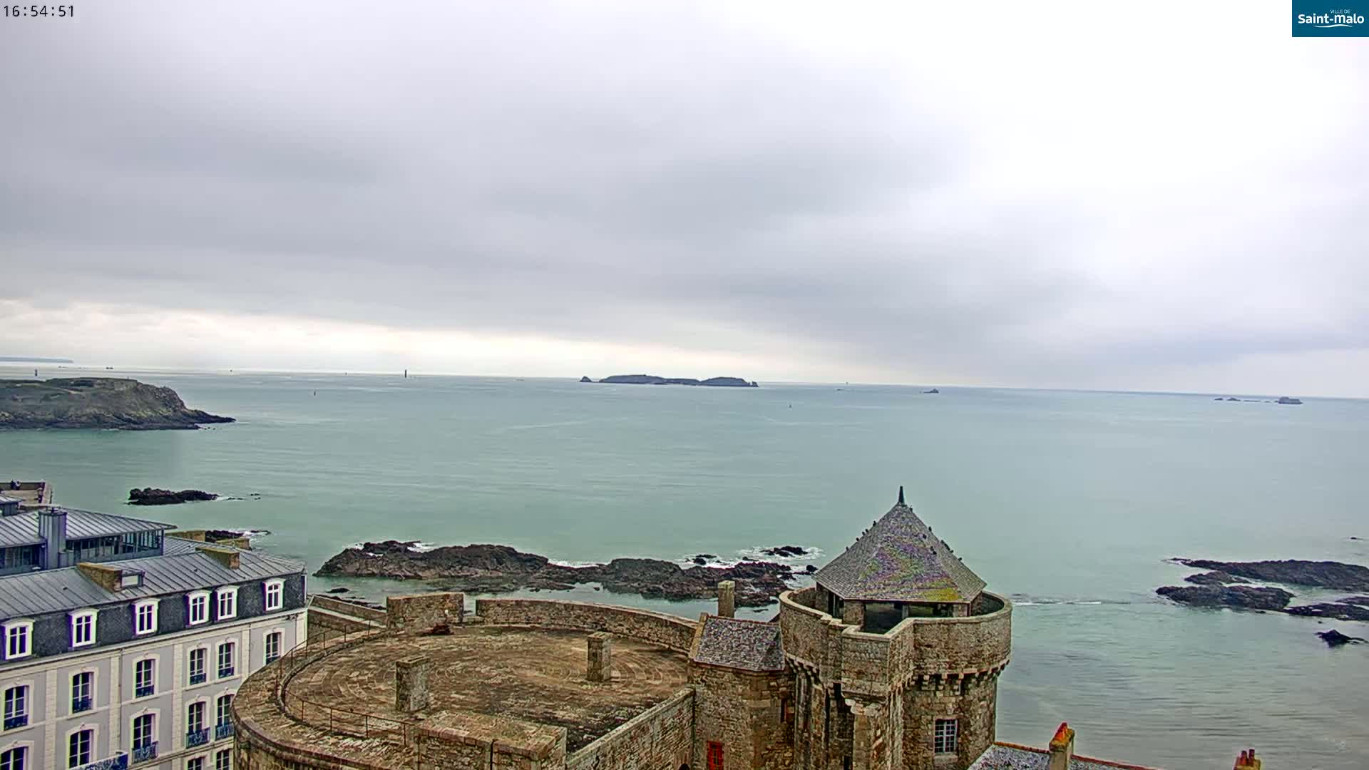 A stone castle overlooks a calm, overcast ocean with rocky coastlines and distant islands.