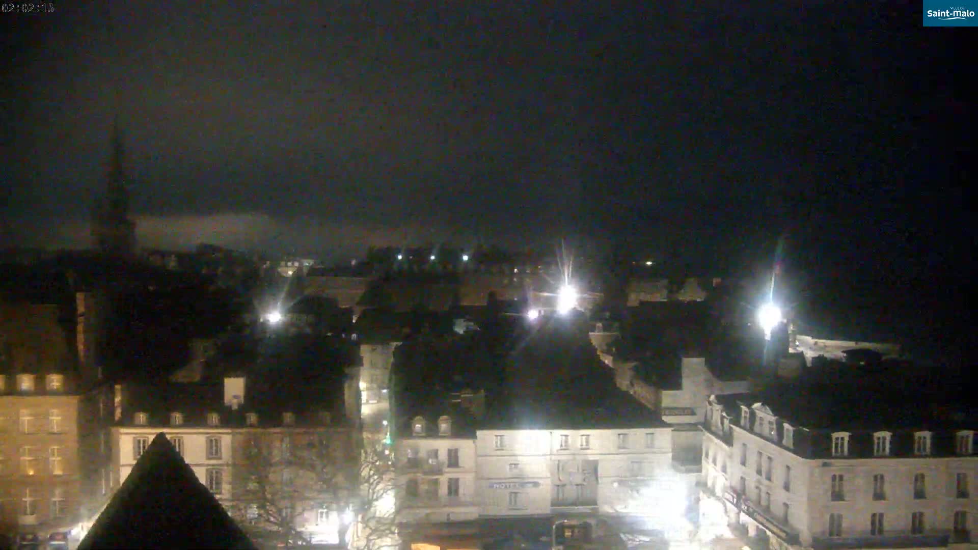 A nighttime view of a city with many buildings lit by streetlights under a dark, cloudy sky.