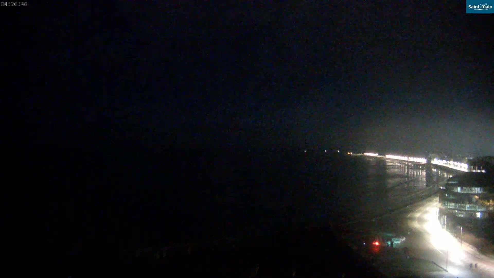 A dark, nighttime view shows a long, illuminated pier extending into a dark body of water, with a road and buildings visible along the shoreline.
