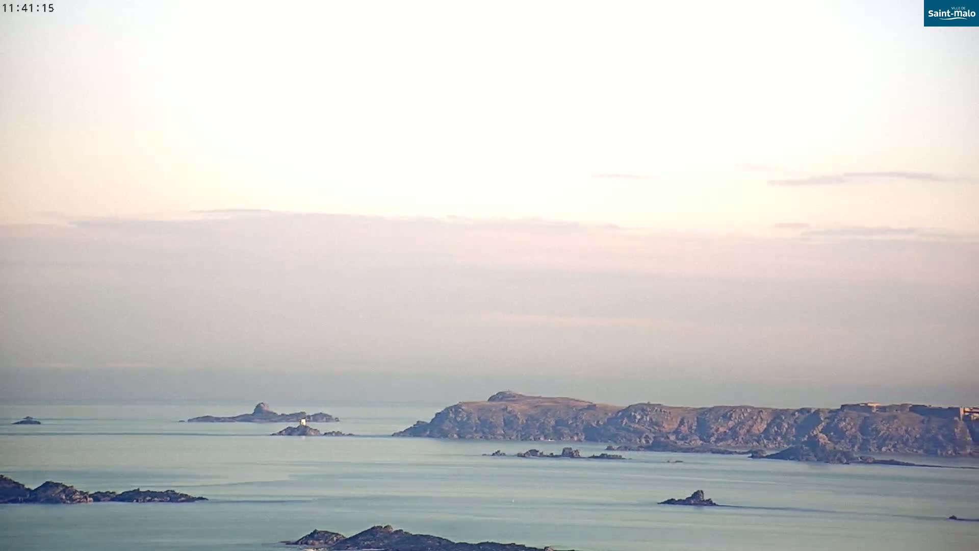 A hazy, calm ocean scene shows several rocky islands and islets under a pale sky.