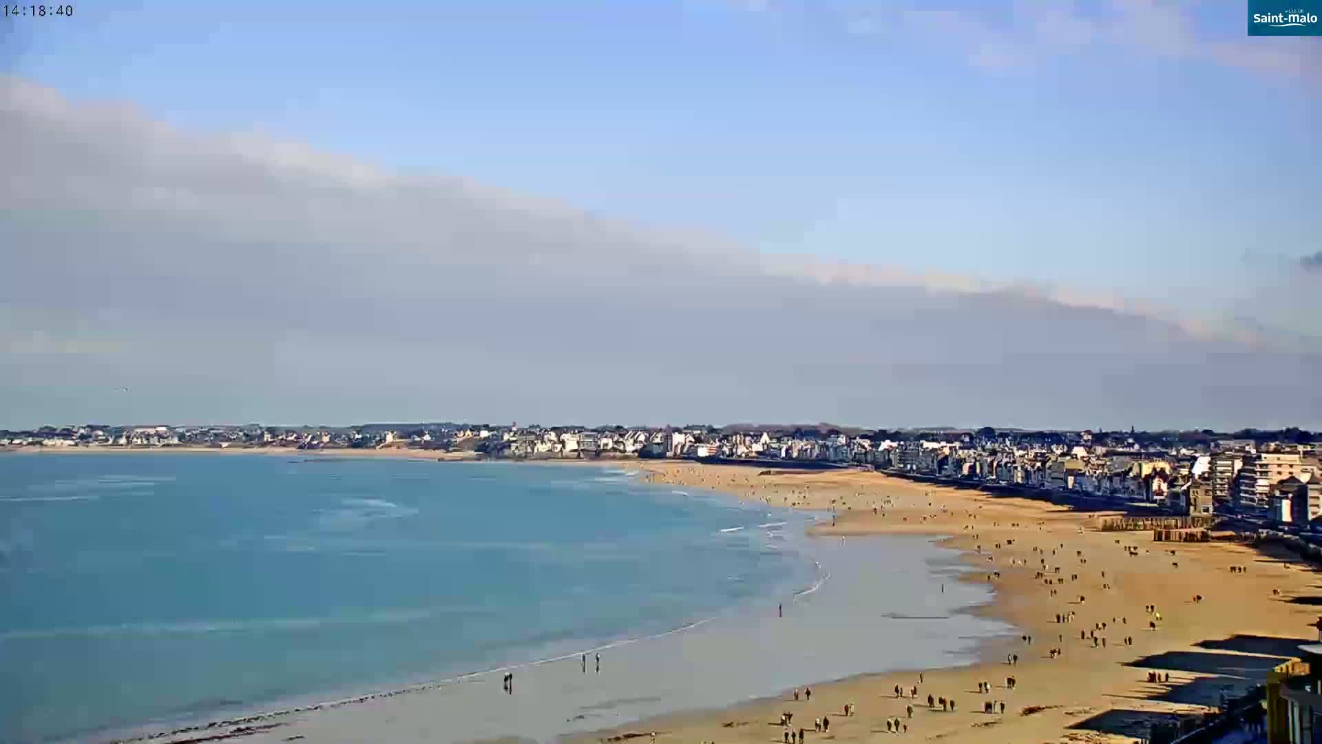 A mostly sunny day shows a long sandy beach with many people on it, curving alongside a calm, light-blue ocean toward a town in the distance.