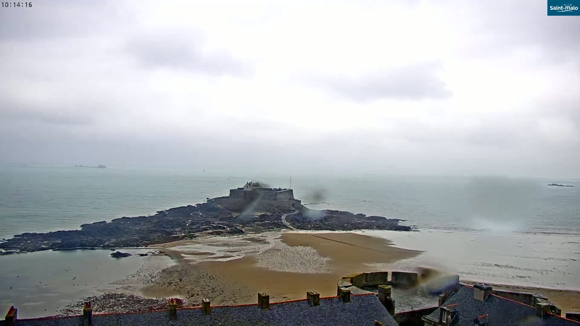 A coastal fortress sits on a rocky island connected to a sandy beach by a narrow strip of land, all under a cloudy, rainy sky.