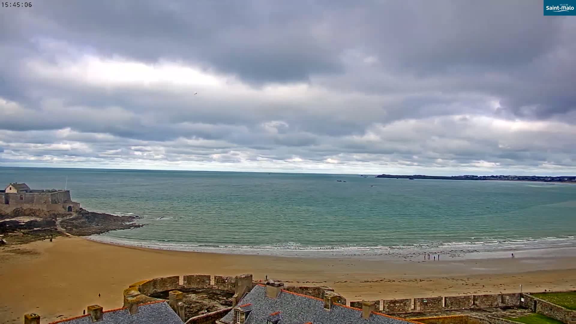 A sandy beach curves around a stone fort next to a calm, teal sea under a mostly cloudy sky.