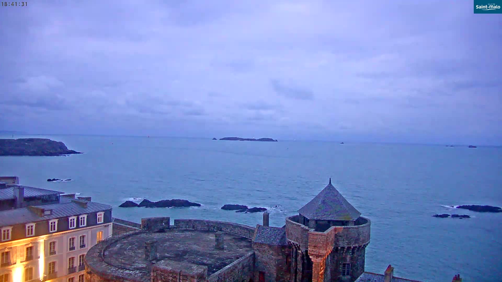 A stone coastal fortress overlooks a calm, twilight sea dotted with rocky islands under a mostly cloudy sky.