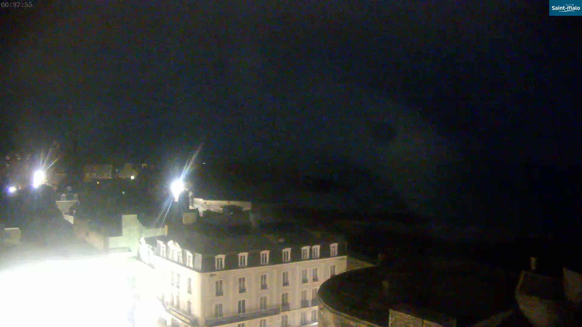 A nighttime, high-angle view of a town with a multi-story building illuminated in the foreground, under a dark sky.