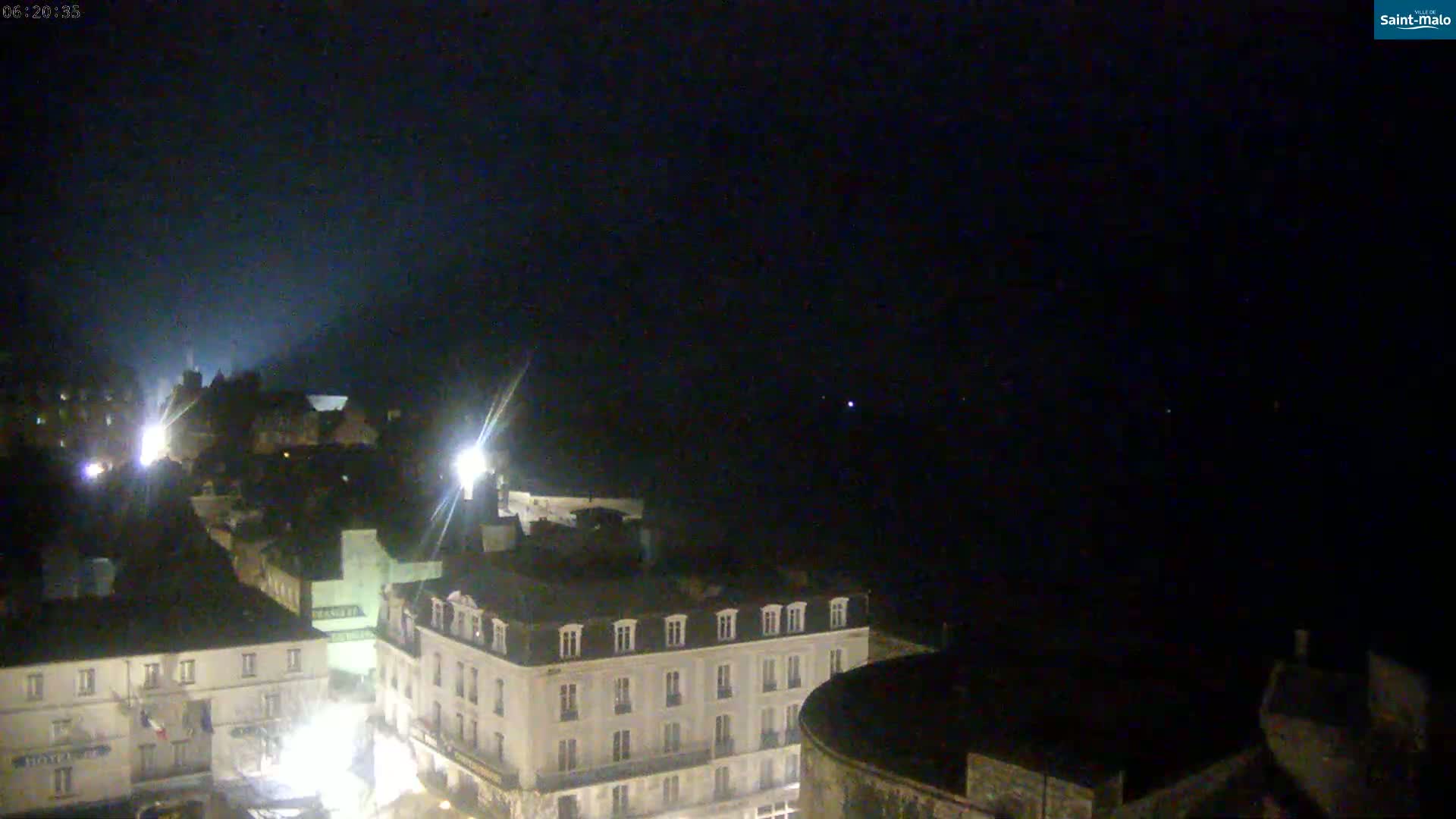 A nighttime, high-angle view shows a European-style town with buildings illuminated by streetlights under a dark, clear sky.