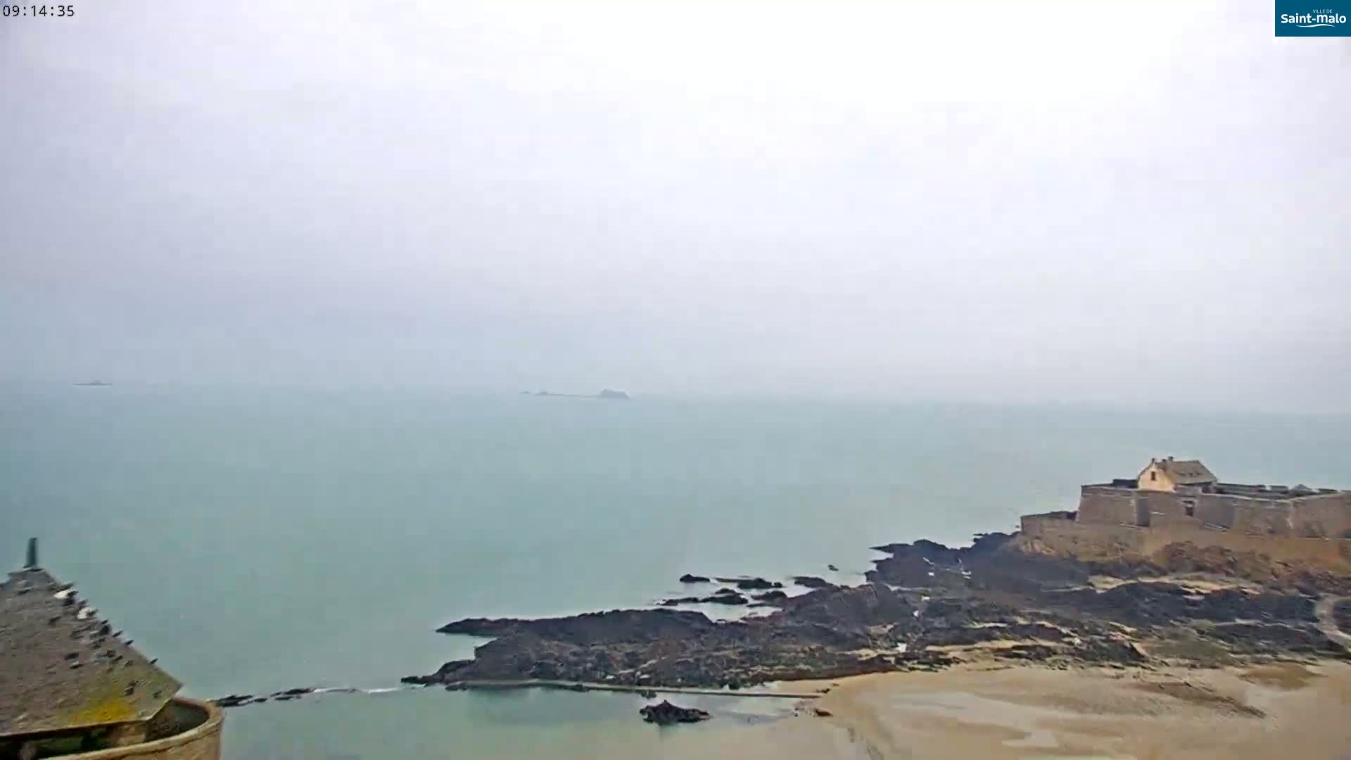 A coastal fortress sits on a rocky outcrop overlooking a hazy, calm sea with a small island visible in the distance.