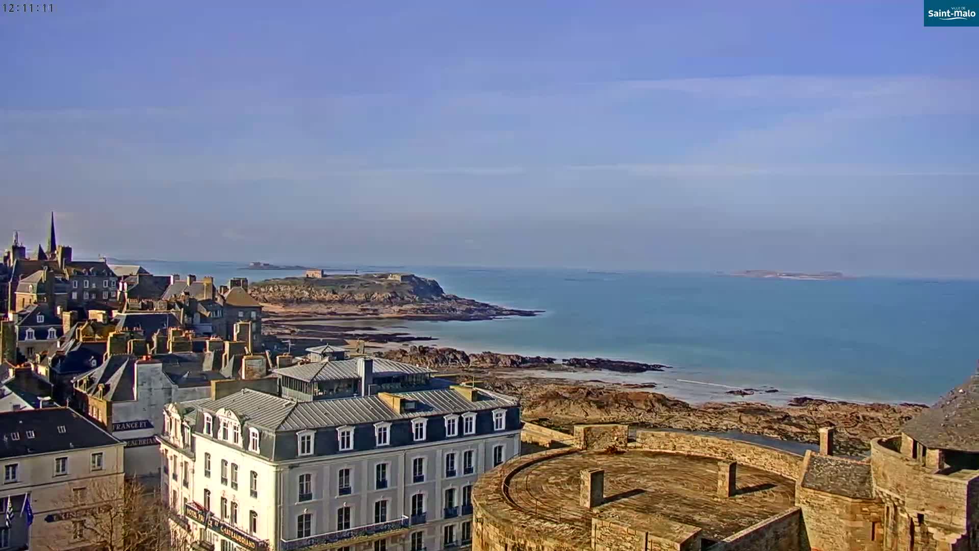 A coastal town with stone buildings and a partially visible castle overlooks a calm, blue ocean under a clear sky.