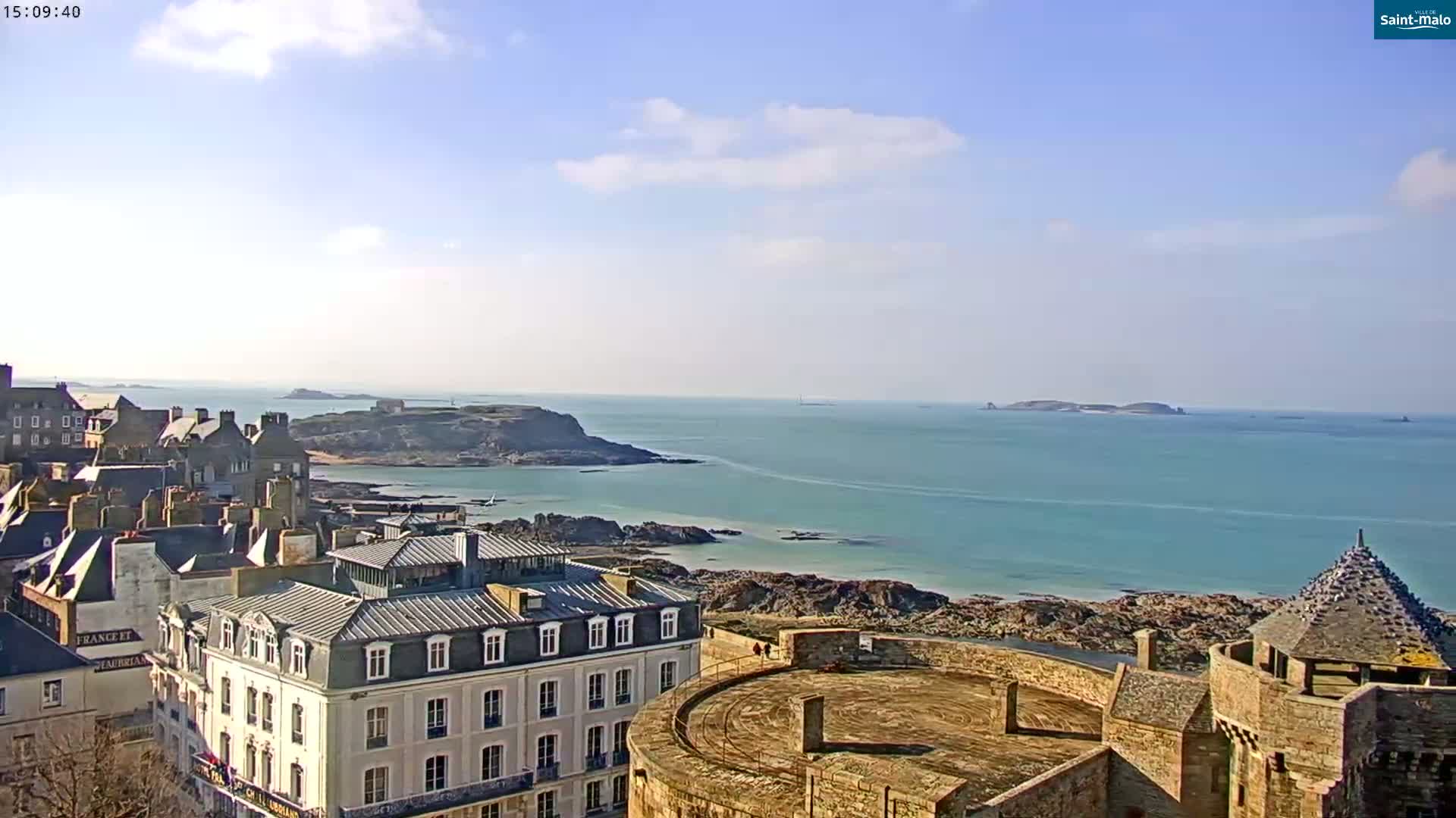 A coastal town with stone buildings overlooks a calm, blue ocean and rocky shoreline under a mostly sunny sky.