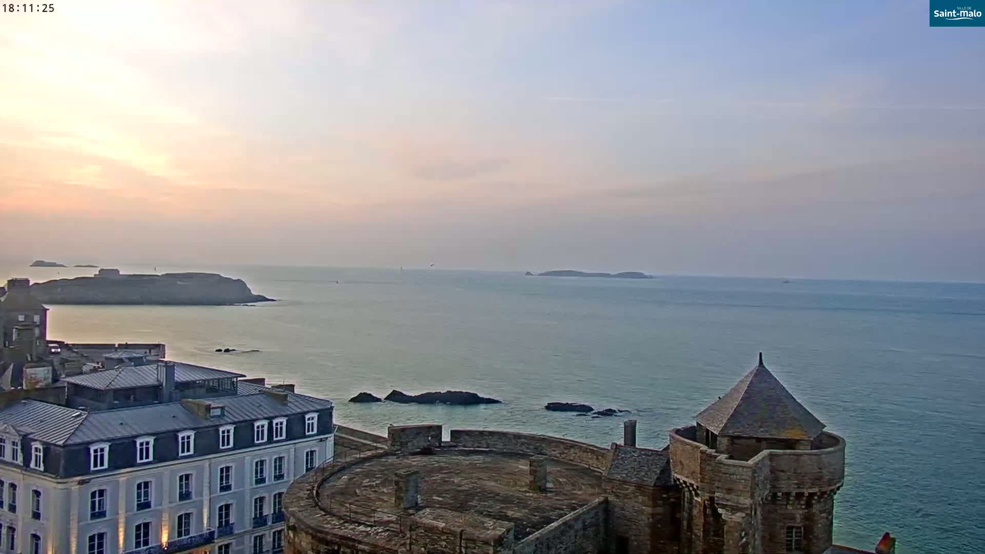 A coastal cityscape at sunset overlooks a calm ocean with distant islands, featuring a stone castle with a conical tower in the foreground.